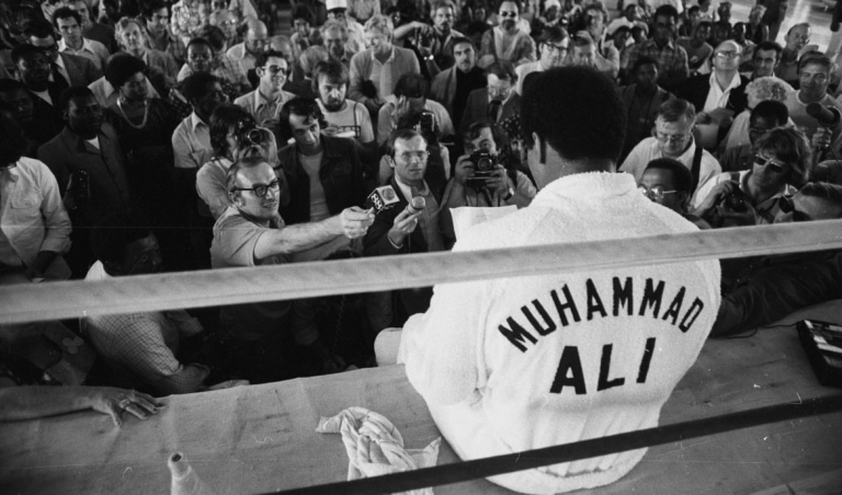 Vintage photo of Muhammad Ali sitting on the outside edge of a boxing ring speaking to a group of reporters