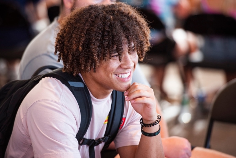 A student wearing a t-shirt and a backpack sits, listens, and smiles