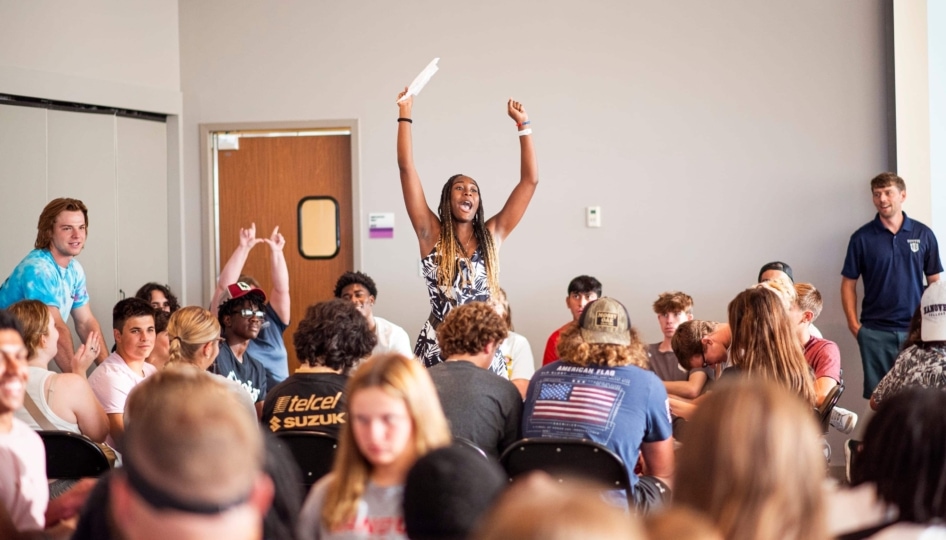 Woman standing with her arms raised high amidst a room full of college students