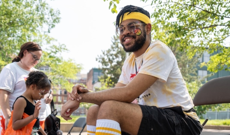 Man sitting in a chair with a headband, glasses, and a peace sign painted on his face with a young child and woman in the background