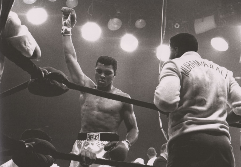 Black and white photo of Ali raising glove in the air in boxing ring after fight