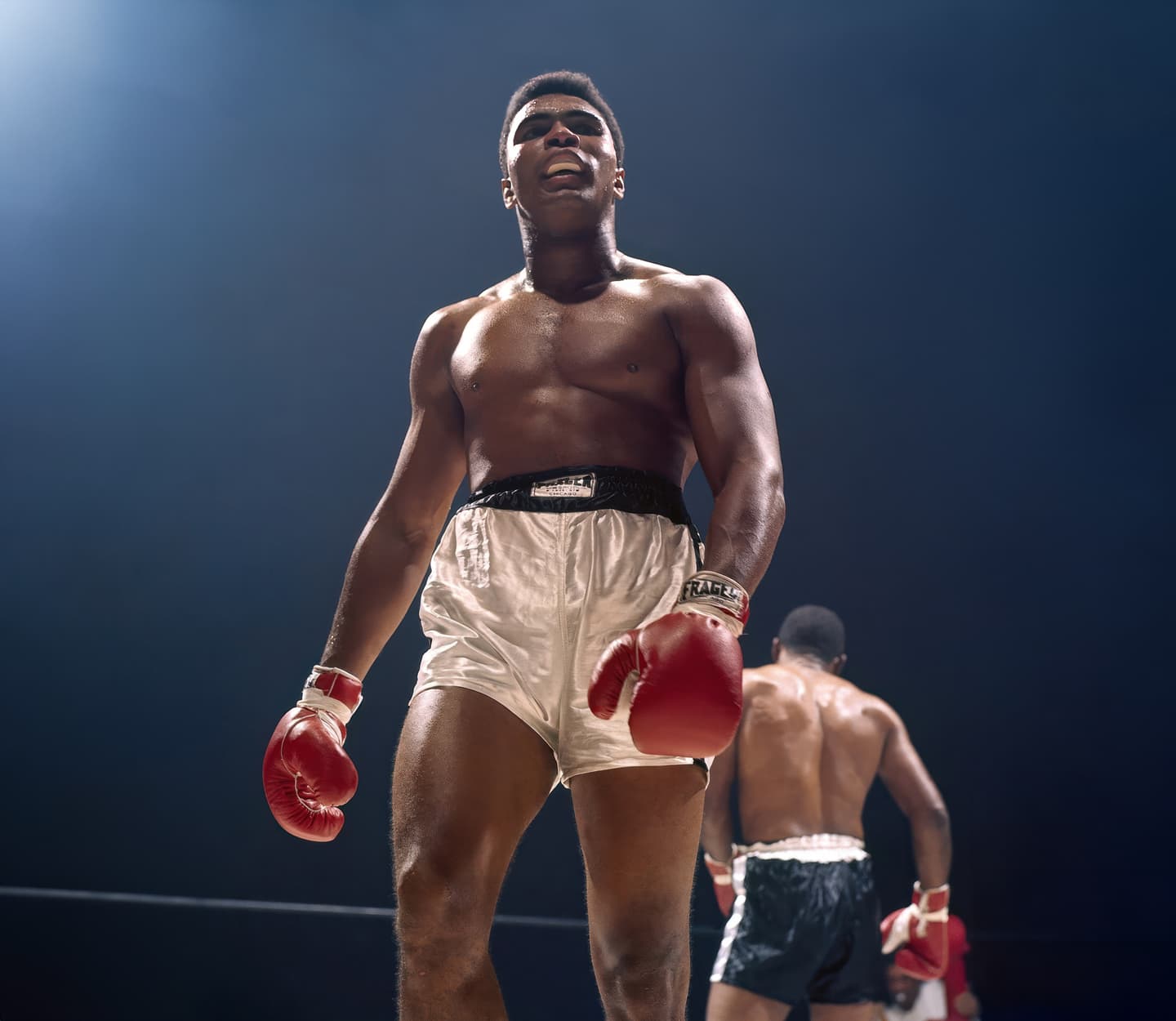 Muhammad Ali standing in a boxing ring in front of another boxer with his back to the camera