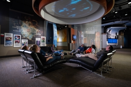 Six museum visitors in an exhibit sitting in reclined chairs looking up at a projection on the ceiling