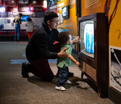 Father and young child kneeling in front of an old television in an exhibit