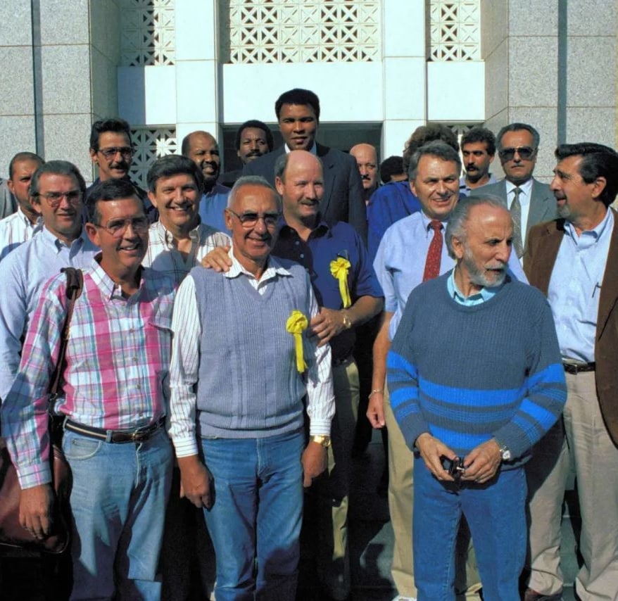 A group of men with yellow ribbon pens on stand in a group. Muhammad Ali is in the back.