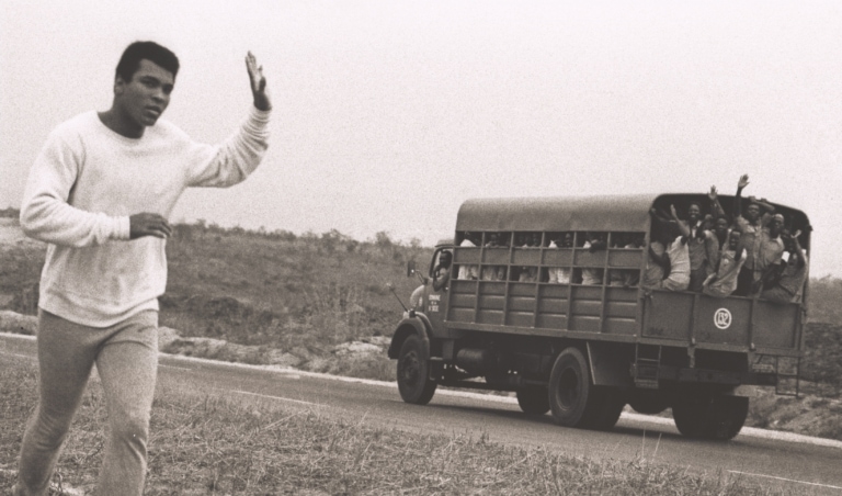 Black and white photo of Muhammad Ali running in sweats while waiving to a large truck full of people waving at him