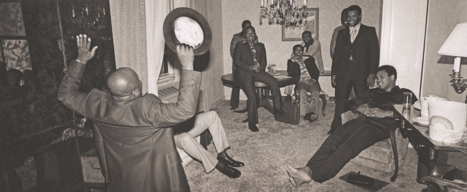 Black and white photo of Muhammad Ali in a home with several men while one man kneels on the floor and gestures wildly with his hat