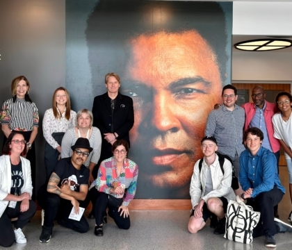 Group of people kneeling and standing in front of a large wall with a photo of Muhammad Ali's face