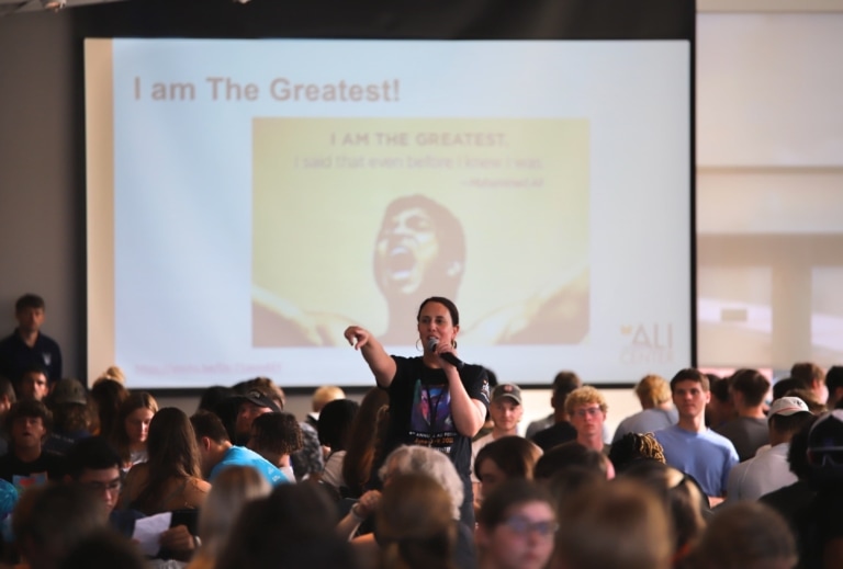 A woman with a microphone is standing in front of a projection screen amongst a room of students pointing to someone or something out of view