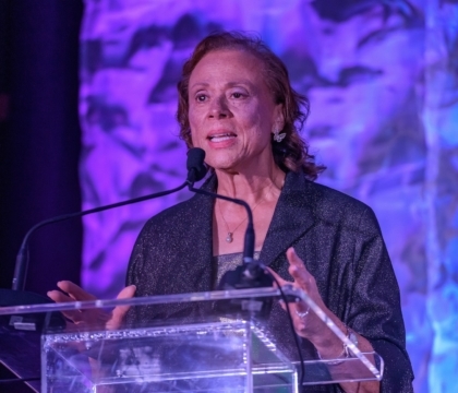 Lonnie Ali speaking on a stage while standing at a clear glass podium in front of a purple background