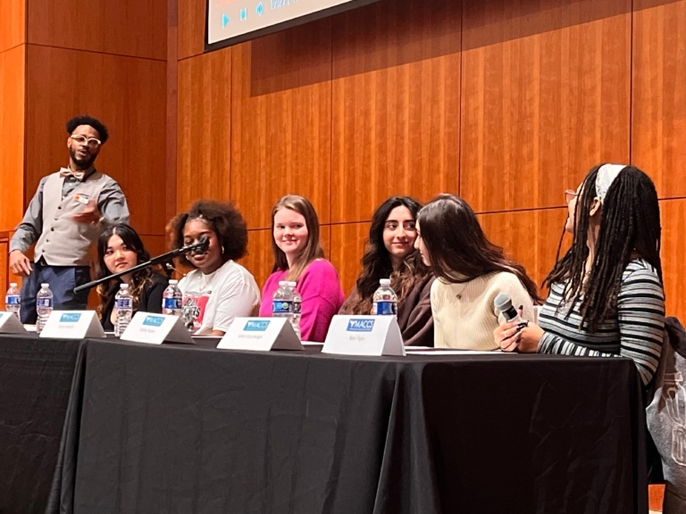 A group of 6 teens sit behind a table with "MACCS" table tags in front of them to give a panel talk while a person in glasses and a bowtie is nearby