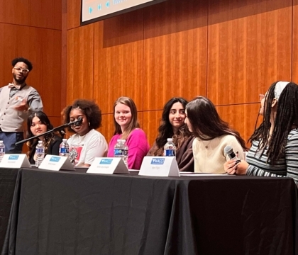 A group of 6 teens sit behind a table with "MACCS" table tags in front of them to give a panel talk while a person in glasses and a bowtie is nearby