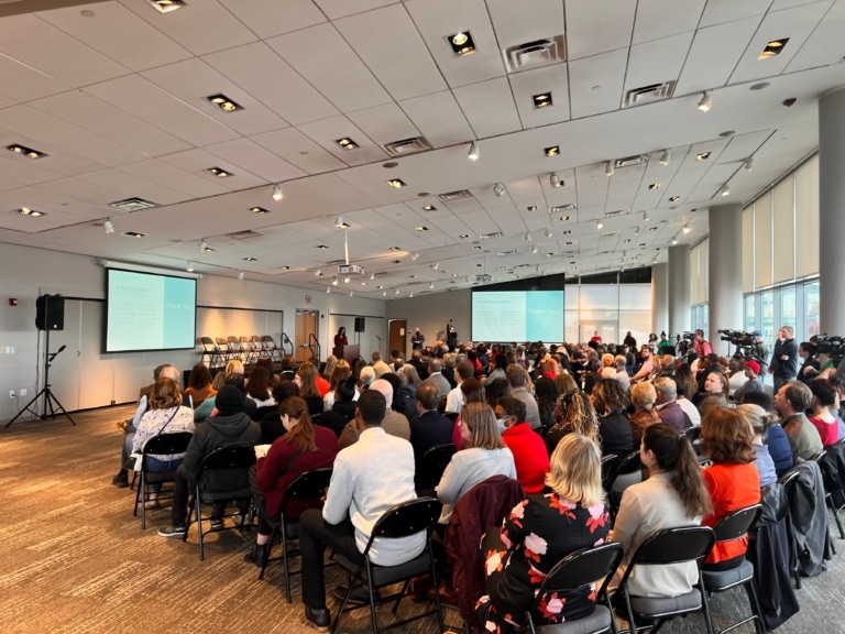 A large group of adults sit in an a large ballroom listening to a person at a lectern speak